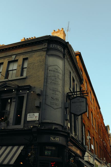 Exterior view of a historic four-story building on a city street with a curved corner, featuring large multi-pane windows and decorative architectural details. The building is painted in light grey and warm orange tones, with a sign reading 'The Marylebone' on the upper portion and a smaller hanging sign indicating 'No. 98.' Adjacent to it, there is a striped awning over the entrance and street signs, including one for 'Moor Street W1.' The scene is illuminated by natural daylight, highlighting the building's textured facade and classic design, typical of Marylebone architecture. This image emphasizes the charming, historic building exterior which complements the focus on professional domestic cleaning and surface maintenance services offered by Marylebone Cleaners.