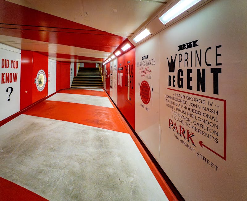 A brightly lit underground corridor featuring walls painted in white and vibrant red, with a smooth, polished concrete floor that appears clean and well-maintained. On the right wall, there are various vintage-style posters and signage, including a notable one for the Prince Regent and historical references, printed in black and red ink. The corridor is illuminated by a row of ceiling lights, creating an evenly bright environment. At the end, a short staircase with wooden steps and metal handrails leads upward. The space exhibits a combination of clean, glossy wall surfaces and a sleek, polished concrete floor, showcasing professional surface cleaning and maintenance, with no visible dust or dirt. Marylebone Cleaners offers expert cleaning solutions to keep such spaces hygienic and visually appealing.